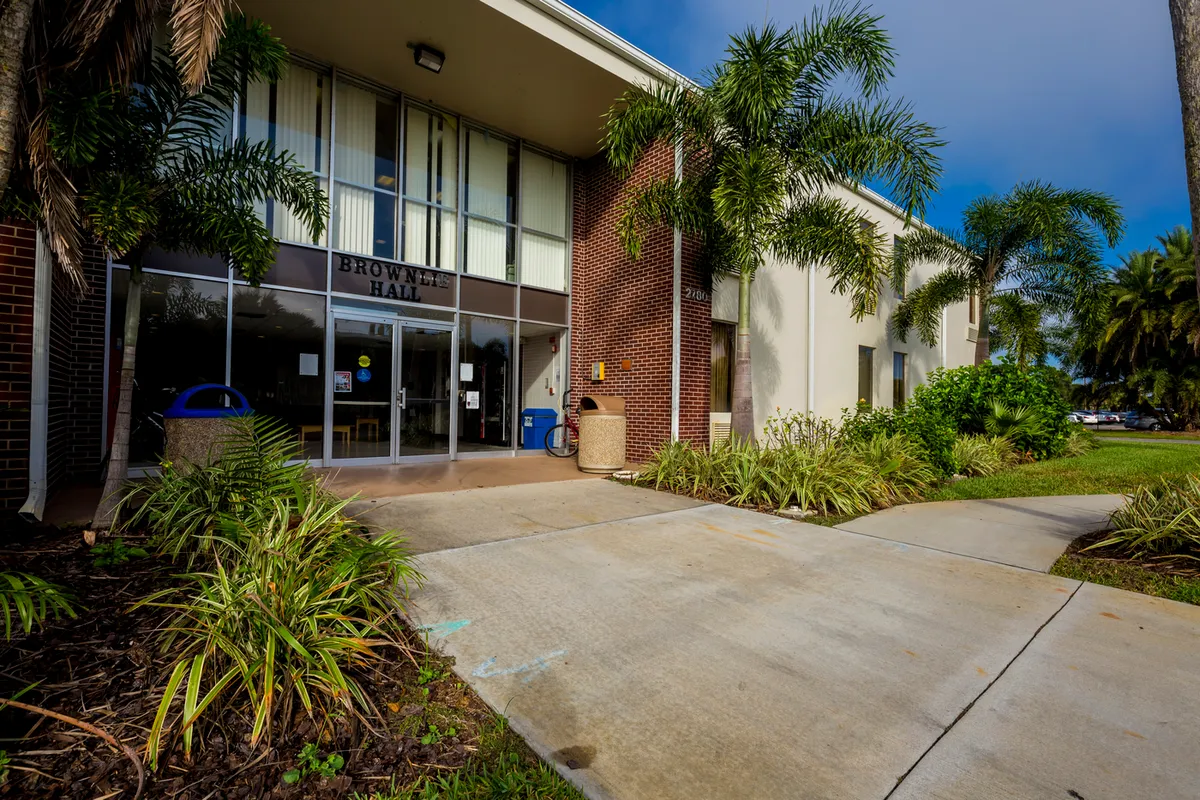 Entryway to two story building, the entryway features two story glass windows and is surrounded by palm trees and tropical plants