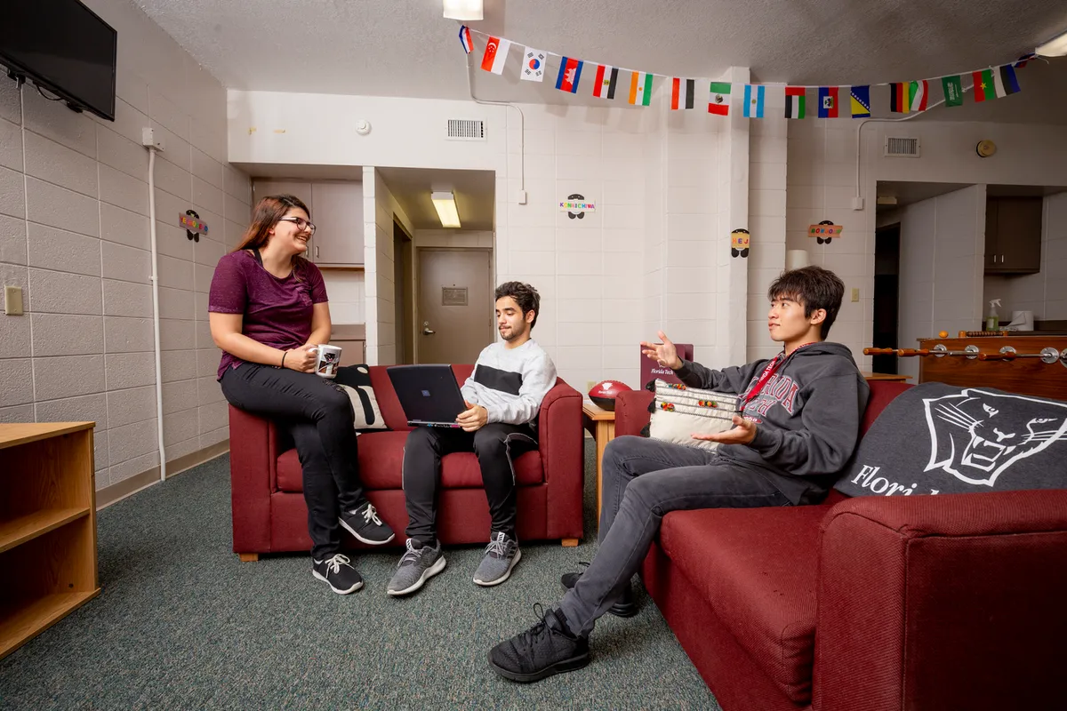 Three students hanging out in a residence hall common area. A TV, foosball table, and couches are visible