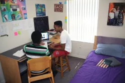 Two students having a conversation around a laptop on a desk in a dorm room. Posters hang on the walls and bed with bright colored bed spread is visible to one side of the room.