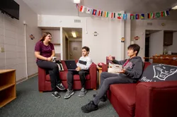 Three students hanging out in a residence hall common area. A TV, foosball table, and couches are visible